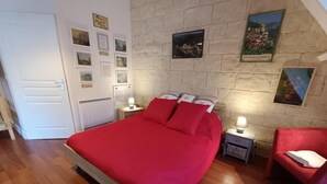 A bedroom with a wooden floor, a bed with red bedding, a nightstand with a lamp, a red chair, and a wall with framed pictures and a stone wall. - Gîtes de France® - My Loft (ST CIRQ SOUILLAGUET)