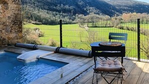 En træterrasse med pool, stenvæg, et bord med bænk og udsigt over en grøn mark og bakker.