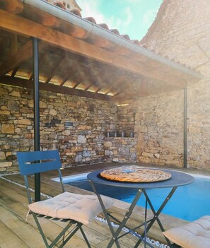 Un espace extérieur entouré de murs en pierre, avec une piscine, une table ronde recouverte d’une nappe tissée et une chaise bleue.