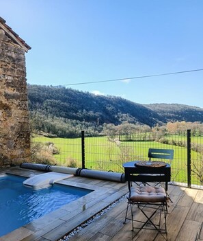 Una terraza de madera con piscina, un muro de piedra, una mesa con banco y vistas a un campo verde y colinas.