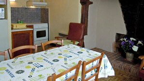 Uma sala de jantar com mesa coberta por toalha estampada com palavras relacionadas a comida, cadeiras de madeira, lareira de tijolos e área de cozinha com fogão e pia.