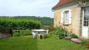 Uma casa com telhado de telhas vermelhas, fachada branca e um pequeno jardim com uma mesa e cadeiras.