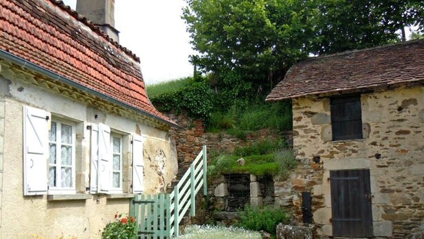 Um edifício rústico de pedra com telhado de telhas vermelhas, uma pequena escadaria que leva a um muro de pedra e um jardim com flores e grama.