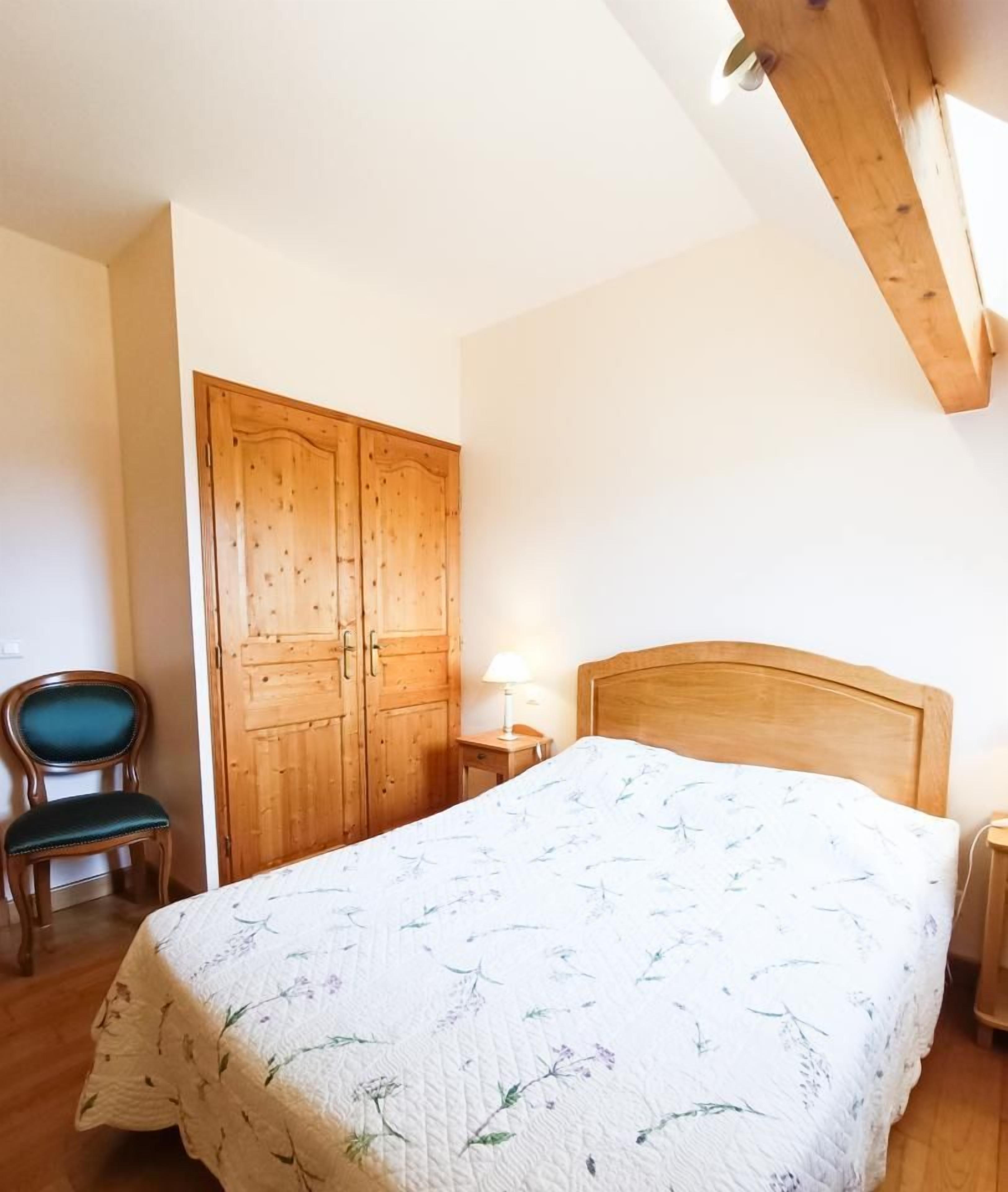A bedroom with a wooden bed frame, a wooden wardrobe, a chair, and a bedside table with a lamp.