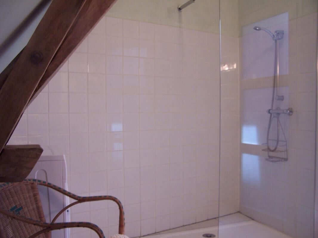 Ein Badezimmer mit Duschkabine aus Glas, weiß gefliesten Wänden und einem Holzbstuhl.