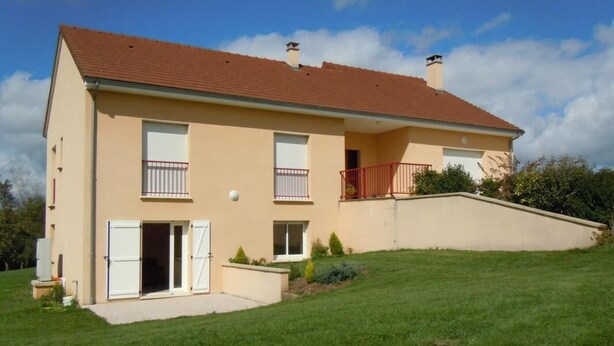 Ein zweistöckiges Haus mit rotem Ziegeldach, einem Balkon mit roten Geländern und einem kleinen Vorgarten mit Rasen und einigen Sträuchern.