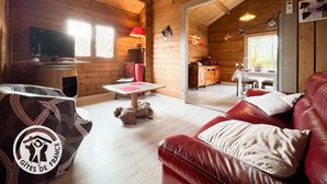 A cozy wooden cabin interior with a leather sofa, a television, a dining table, and a small dog. - Gîtes de France® - My Little Paradise (CARNAC ROUFFIAC)