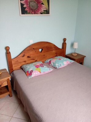 Un dormitorio con cabecera de madera, dos almohadas con estampado floral, una mesita de noche con lámpara y un cuadro enmarcado en la pared.