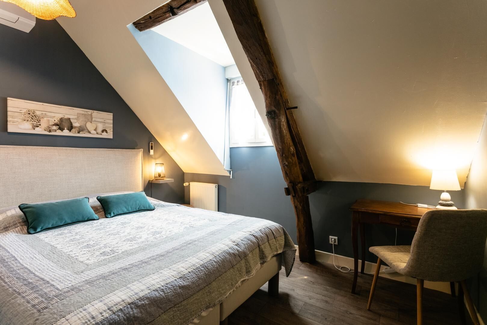 Une chambre avec un plafond en pente, un lit recouvert d’une couvre-lit à motifs, deux oreillers, une table de chevet avec une lampe, une chaise et un tableau accroché au mur.