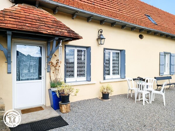 A building with a tiled roof, a door, and windows with shutters. There are potted plants and outdoor furniture.