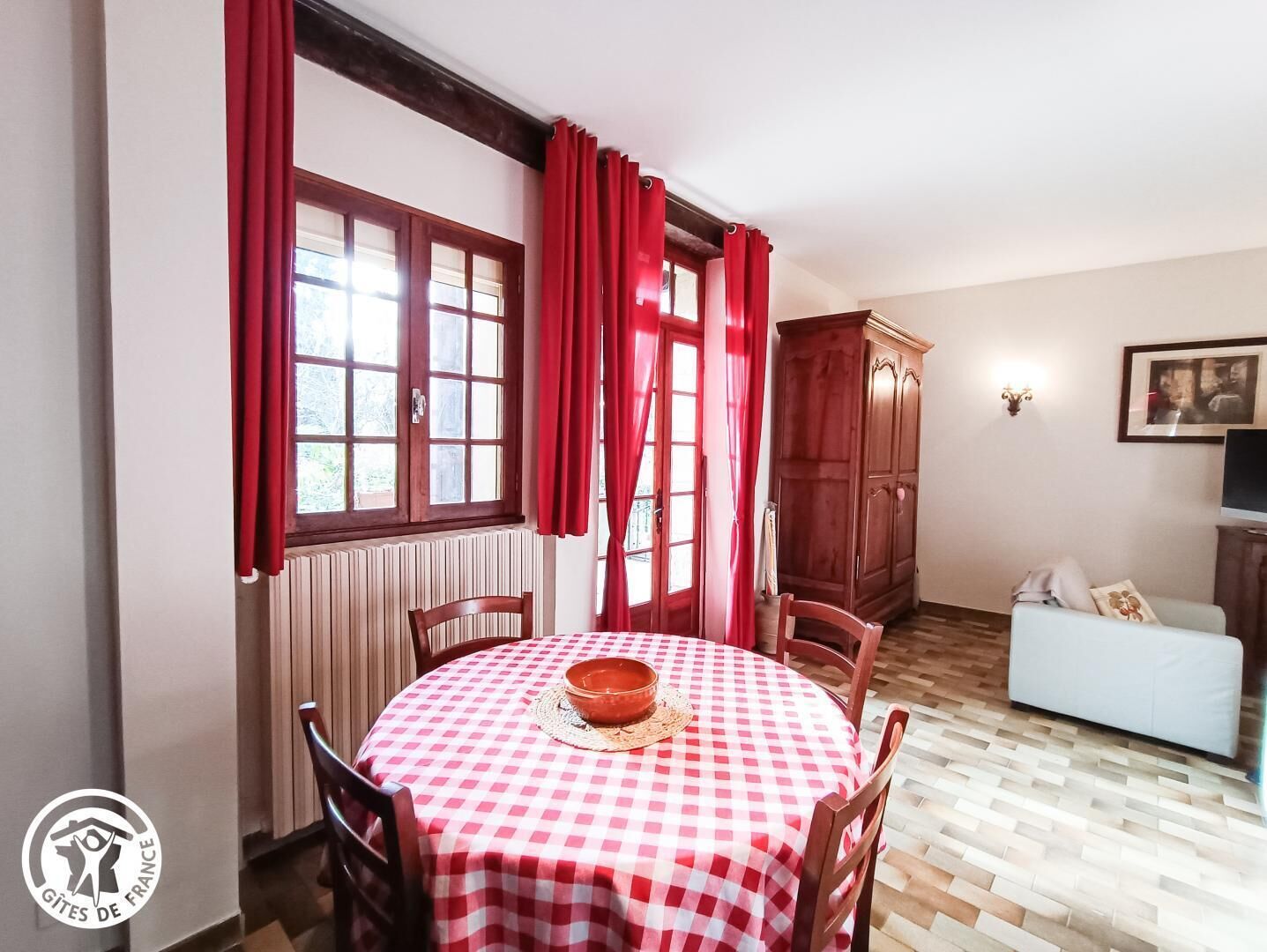 Un comedor con una mesa redonda cubierta por un mantel a cuadros rojo y blanco, sillas de madera y un armario de madera.