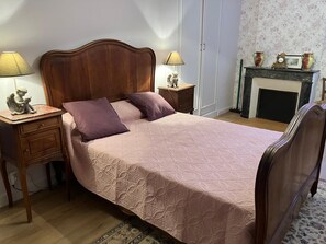 Una cama bien hecha con cabecera de madera, dos mesitas de noche con lámparas, una pequeña estatua y una chimenea con un reloj y un marco de fotos encima.