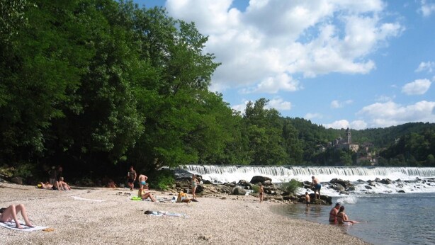 Una playa de guijarros con gente tomando el sol y nadando, una cascada al fondo y una iglesia en una colina.