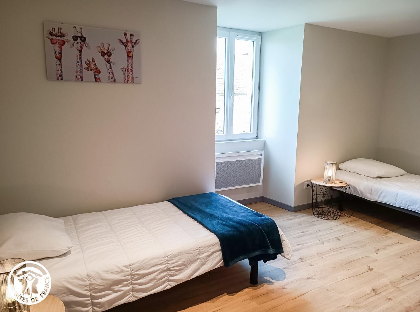 Een slaapkamer met een bed, een bijzetbed, een nachtkastje, een raam en een schilderij van giraffen aan de muur.