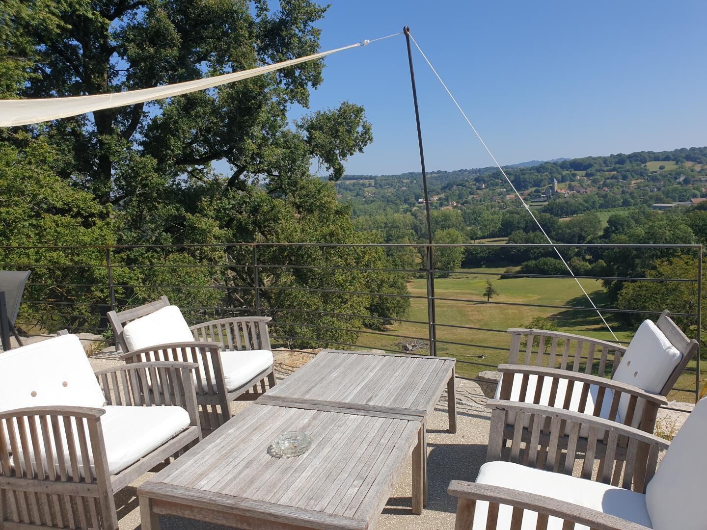 An outdoor seating area with wooden tables and chairs, a canopy, and a view of a green landscape with trees and hills.