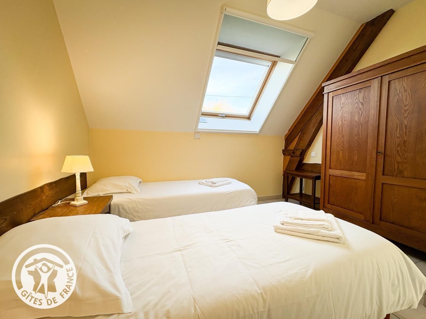 Une chambre avec deux lits, une lucarne, une armoire en bois et une table de chevet avec une lampe.