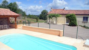 Una piscina con cubierta de baldosas, zona de estar techada y jardín cercado.