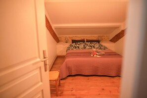Un dormitorio con techo inclinado, una cama con cabecera estampada con motivos florales, una mesita de noche pequeña con lámpara y una silla de madera.