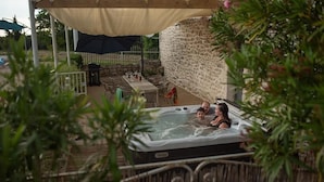 Un jacuzzi con tres personas bajo una cubierta, rodeado de muebles de exterior y un muro de piedra.