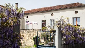 Un bâtiment avec un mur en pierre, un portail en métal et des plantes grimpantes aux fleurs violettes.