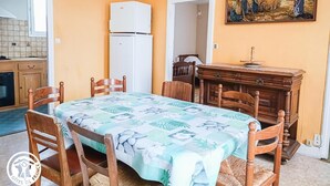 Un coin repas avec une table dressée pour quatre personnes, entourée de chaises en bois. Un réfrigérateur blanc est adossé au mur, et un tableau est accroché au-dessus de la table.