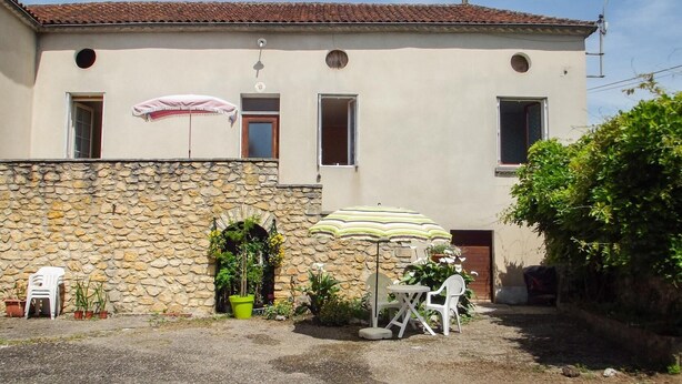 Un bâtiment doté d’une façade en pierre, d’un toit recouvert de tuiles et d’un petit patio avec une table et des chaises placées sous un parasol à rayures.