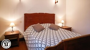 A bedroom with a wooden headboard, a patterned bedspread, two bedside lamps, and a small wooden nightstand.