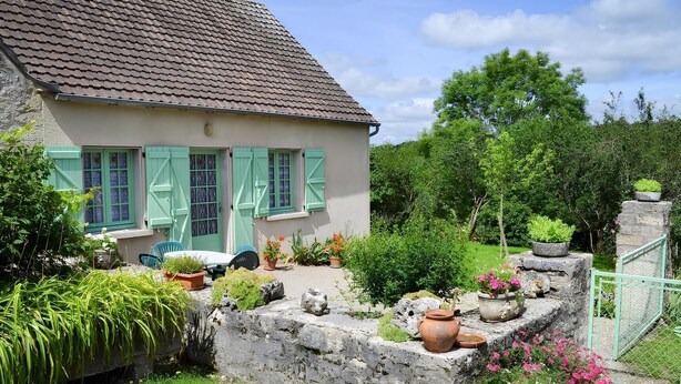 A house with a tiled roof, green shutters, and a garden with potted plants and flowers.