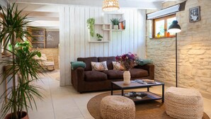 Ein gemütliches Wohnzimmer mit einem braunen Sofa, einem Couchtisch und zwei Poufs. Es gibt Pflanzen, ein Bücherregal und eine Steinwand.