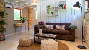 Ein gemütliches Wohnzimmer mit einem braunen Sofa, zwei Poufs, einem Couchtisch und einer Stehlampe. Im Hintergrund befinden sich Blumentöpfe und ein Bücherregal.