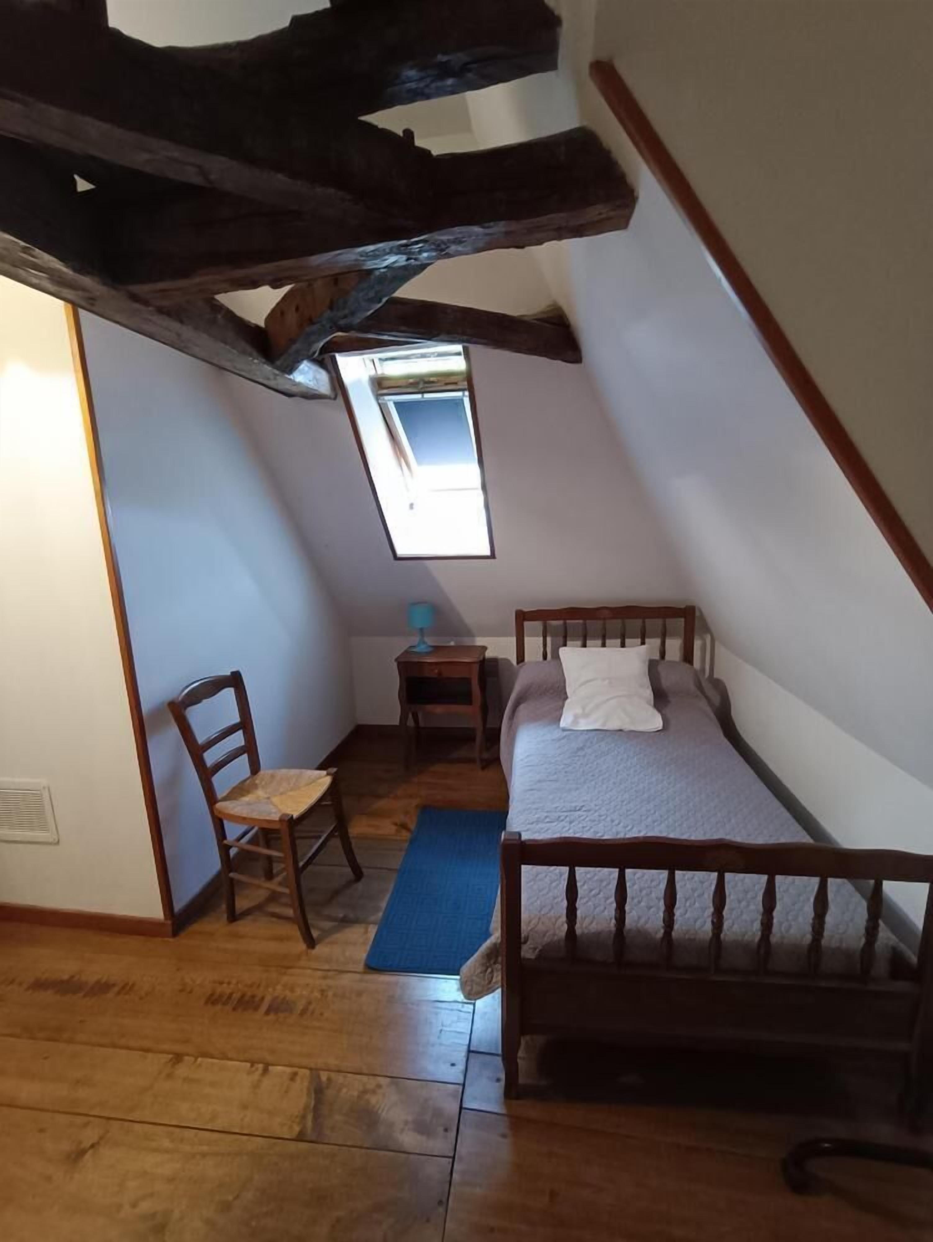 Un dormitorio pequeño estilo ático con techo inclinado, cama individual, una silla, una mesita de noche y un tragaluz.