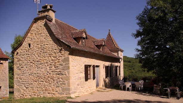 Ein Steinhaus mit Satteldach, Schornstein und einer kleinen Veranda mit Stühlen.