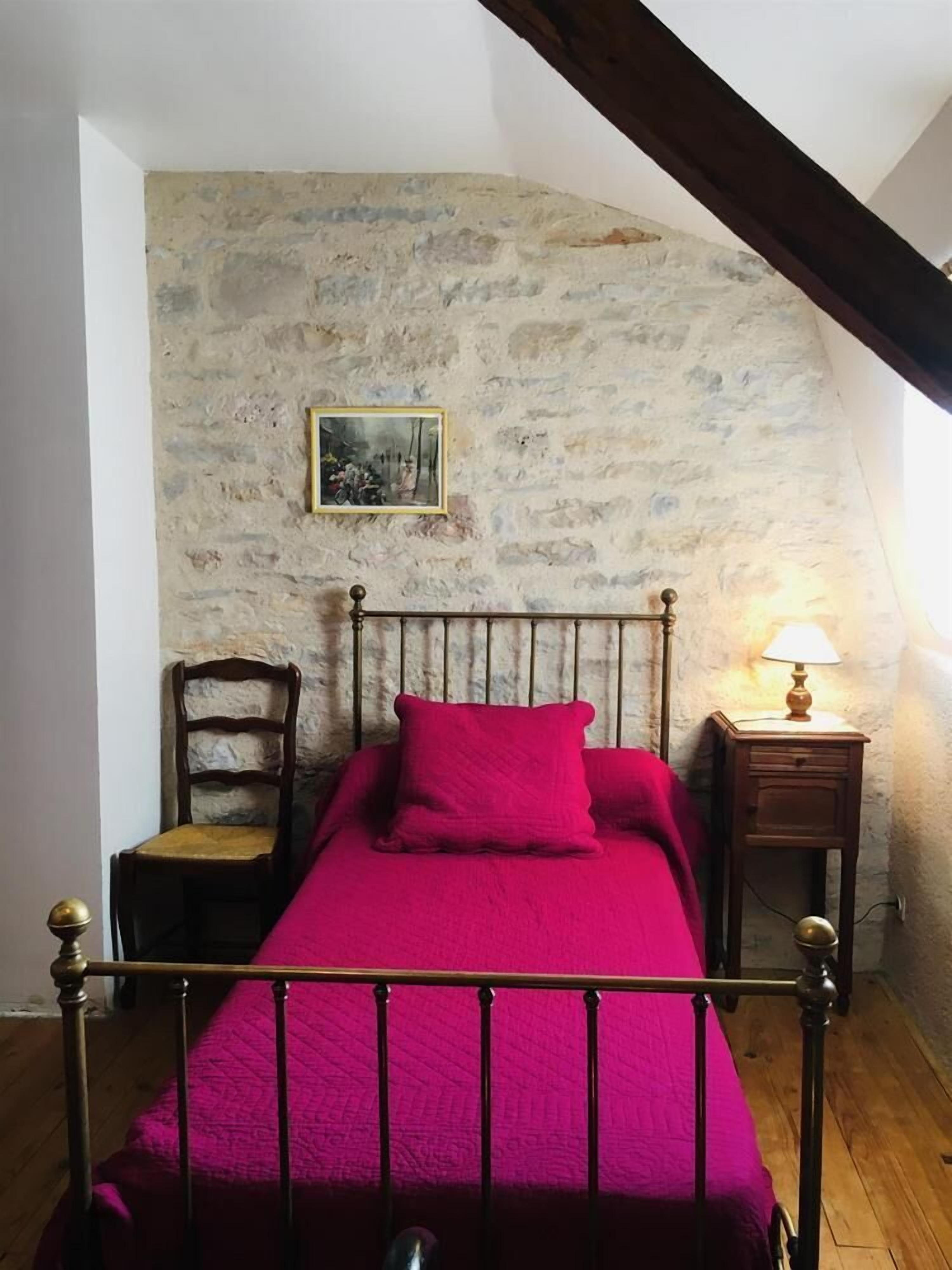 A bedroom with a stone wall, a bed with a metal frame, a wooden chair, a nightstand with a lamp, and a picture on the wall.