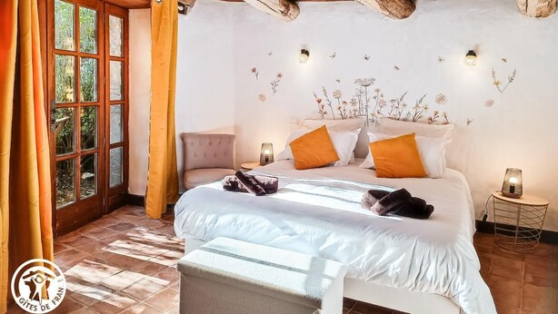 A bedroom with a wooden ceiling, a bed with white bedding and orange pillows, a chair, a small table with a lamp, and a window with curtains.