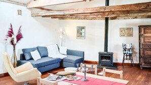 A cozy living room with a wooden ceiling, a blue sofa, a wooden dining table with chairs, a vase with dried flowers, and a small fireplace.