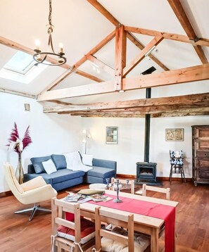 Una sala de estar acogedora con techo de madera, un sofá azul, una mesa de comedor de madera con sillas, un jarrón con flores secas y una pequeña chimenea.