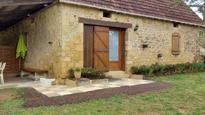 Un edificio rustico in pietra con una porta e persiane in legno, una piccola zona esterna con sedie e tavolo di plastica, e un vialetto lastricato che conduce all'ingresso.
