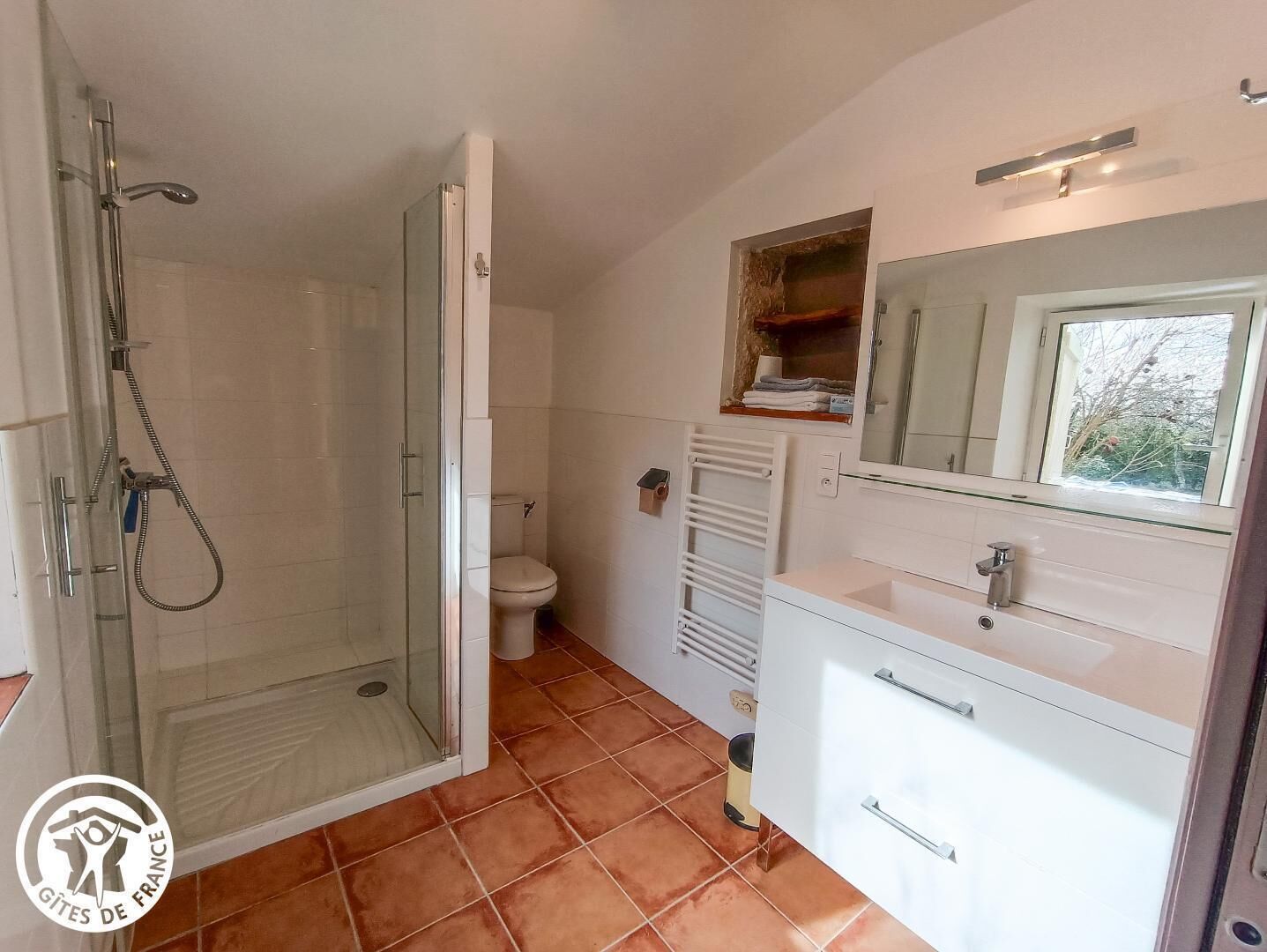 Et badeværelse med glasvæg omkring brusebad, toilet, vask med spejl og håndklædestang.
