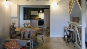 Uma área de refeições com uma mesa redonda de madeira e cadeiras, uma cozinha ao fundo e um espaço bar com banquetas.