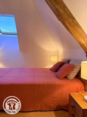 A bedroom with a sloped ceiling, a bed with a red bedspread, patterned pillows, a nightstand with a lamp, and a window with a view of greenery. - Gîtes de France® - Laginestière Guesthouse (CREGOLS)