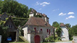Un edificio de piedra tradicional con una puerta roja y un pequeño balcón, rodeado de abundante vegetación y otras construcciones rústicas.