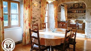 A dining area with a round table, wooden chairs, and floral curtains. There is a window with a view of greenery outside. - Gîtes de France® - Lou Sécadou (CUZAC)