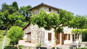 A stone house with a tiled roof, surrounded by greenery and a small patio area. - Gîtes de France® - Lou Sécadou (CUZAC)