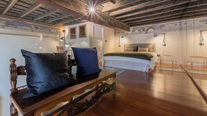 Una habitación con techo de madera, una cama con colcha blanca y verde, una mesa de madera con dos cojines azules y un separador de vidrio.