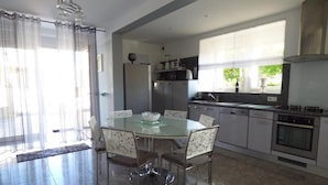 A modern kitchen with a dining area, stainless steel appliances, and a large window providing natural light. - Gîtes de France® - LE PATIO DU BARRY (SAINT-VINCENT-RIVE-D'OLT)