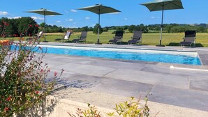 Une piscine entourée de verdure, avec des chaises longues et des parasols, sous un ciel dégagé.
