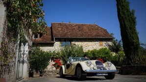 A vintage car parked in front of a stone house with a tiled roof, surrounded by greenery and flowers.