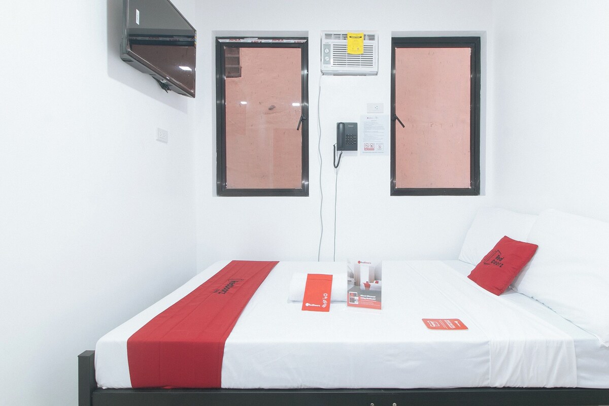 A hotel room with a bed, two windows, an air conditioning unit, and a TV mounted on the wall.