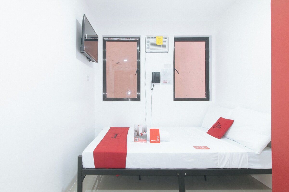 A single-bed room with a white bed, red pillows, a red bedside table, a flat-screen TV mounted on the wall, and a window with a pinkish tint.