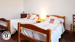 Quarto com duas camas de madeira, cada uma com mesa de cabeceira e luminária. Paredes brancas e piso de madeira.